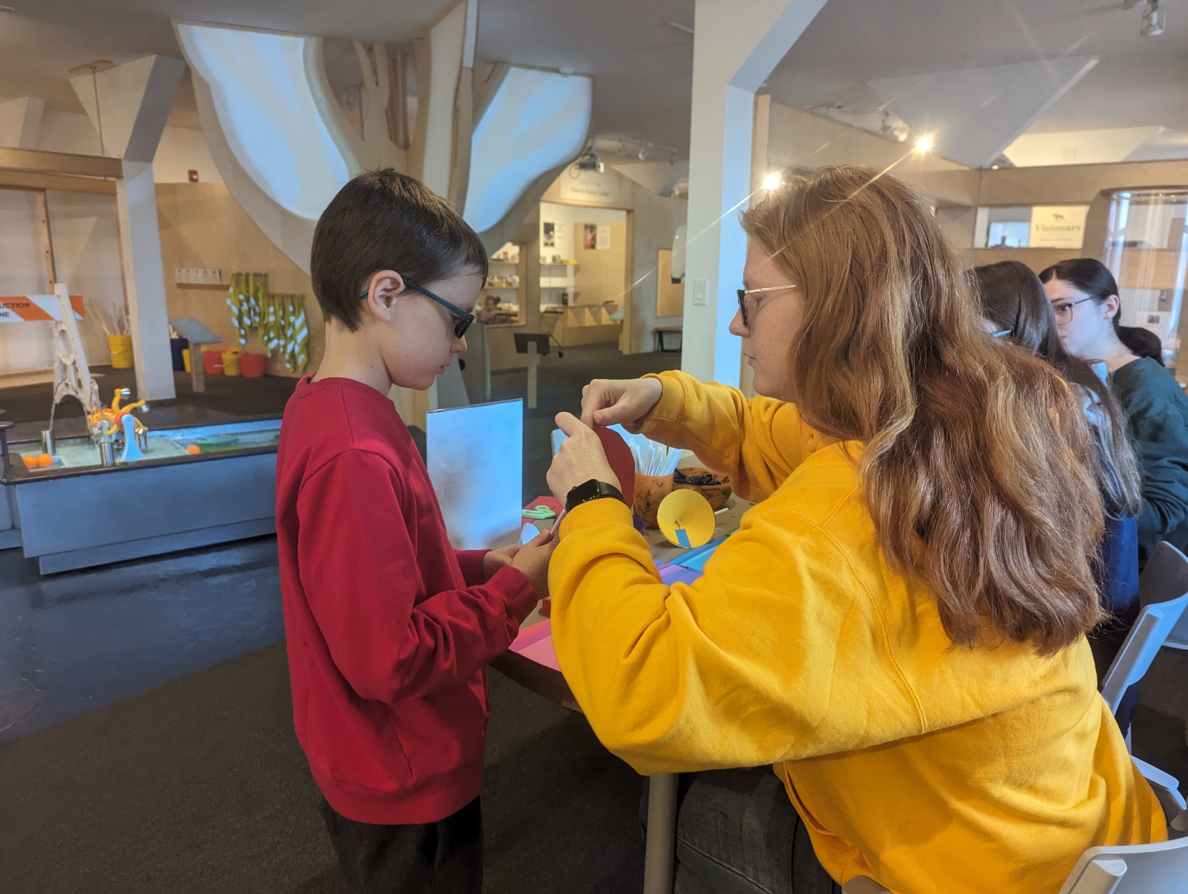 A Clarkson University Honors student wearing a yellow sweatshirt helps a child in a red shirt with a hands-on craft activity at the North Country Children’s Museum. Colorful craft supplies cover the table, with museum exhibits and other participants visible in the background.