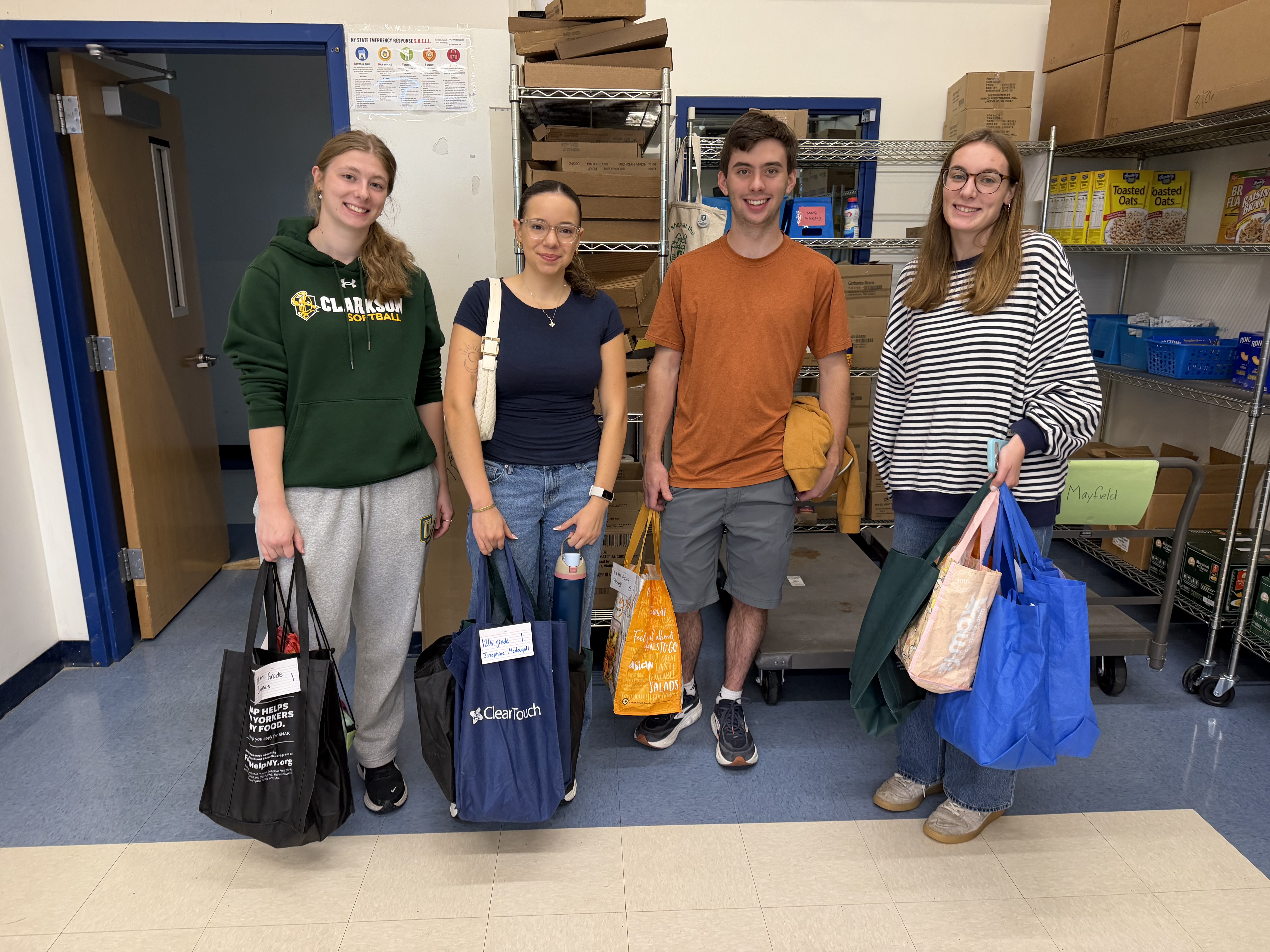 Four Clarkson University Honors students stand together holding reusable bags filled with donated items for the Potsdam Snack Pack Program. Behind them are shelves stocked with food, supplies and cardboard boxes in the program’s storage room.