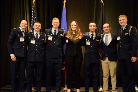 Group of seven people pose together at a formal event, including five individuals in military uniforms and two in civilian attire, with flags displayed behind them.